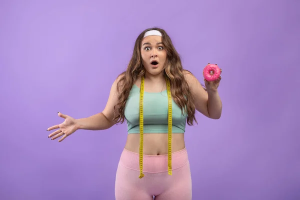 Shocked sad young european plus size lady in uniform with open mouth and measuring tape hold donut