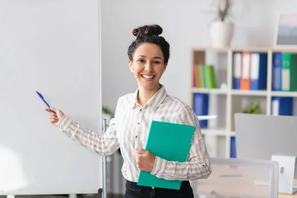 Concepto de presentación. Feliz manager femenino sosteniendo el bloc de notas y apuntando al espacio libre en la pizarra vacía en la oficina —  Fotos de Stock