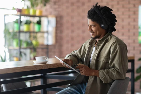 Glada unga afrikanska amerikanska lockigt man i trådlösa hörlurar titta på surfplatta, sitta vid bordet med kopp kaffe i café — Stockfoto