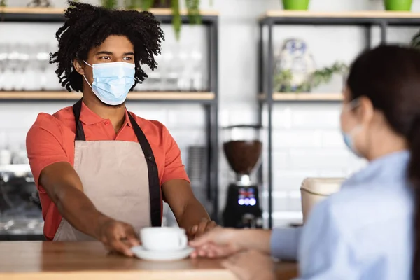 Smiling young african american curly male waiter in protective mask and apron giving fresh coffee to client — Stock Photo, Image