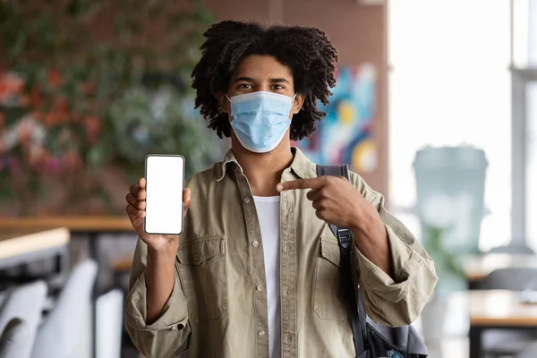 Happy millennial african american curly guy student in protective mask with backpack pointing finger at smartphone — Stock Photo, Image