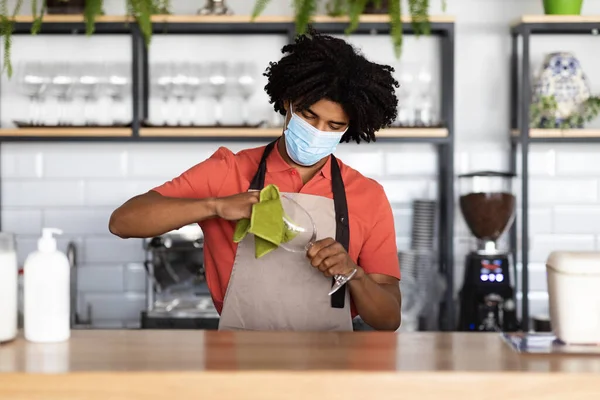 Ocupado millennial preto encaracolado garçom masculino em máscara protetora e avental limpa vidro com pano atrás bar — Fotografia de Stock