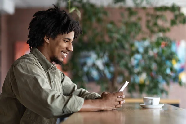 Alegre millennial afroamericano rizado chico blogger escribiendo en el teléfono inteligente en la mesa con una taza de café —  Fotos de Stock