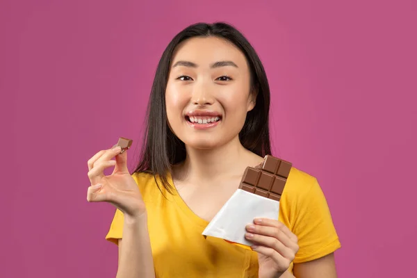 Feliz dama coreana comiendo chocolate, disfrutando de delicioso postre y sonriendo a la cámara, de pie sobre el fondo del estudio violeta —  Fotos de Stock