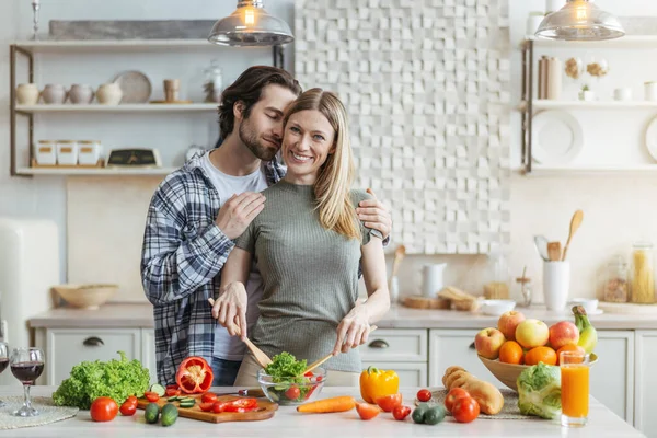 Sorrindo jovem mulher europeia prepara salada, homem com abraços de restolho esposa à mesa com legumes orgânicos — Fotografia de Stock