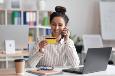 Ofiste dizüstü bilgisayarda çalışan neşeli bir kadın, elinde kredi kartı olan, banka ya da web mağazası müdürünü arayan, cep telefonu kullanan.