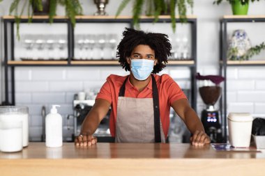 Mutlu, kendinden emin, Afro-Amerikan kıvırcık saçlı, koruyucu maskeli garson, kafe barında önlük satan adam.