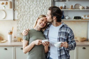 Tatmin olmuş, beyaz, sakallı genç bir adam karısını alnından öpüyor. Mutfağın iç kısımlarında kahve içiyor.