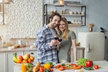 Neşeli genç Avrupalı koca, sakallı, karısına sarılmış, kadeh tokuşturmuş, yemek hazırlıyor.