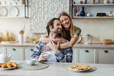 Glad young european lady hugging male with stubble drink coffee, enjoy free time in modern kitchen interior clipart