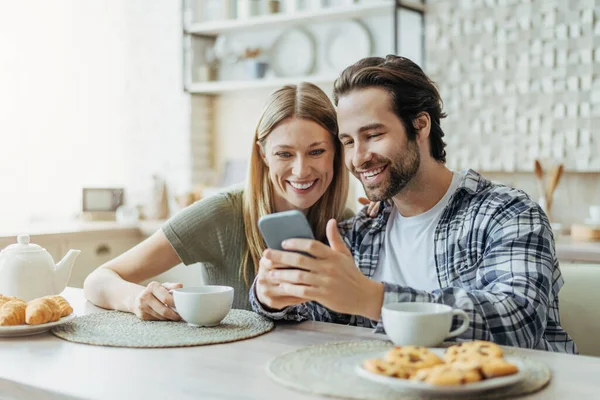 Gülümseyen Avrupalı genç aile akıllı telefona bakar, sosyal ağlarda sohbet eder, modern mutfakta çay içer. — Stok fotoğraf