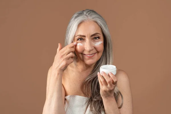 Beautiful senior woman applying moisturizing or nourishing cream under her eyes on brown studio background — стоковое фото