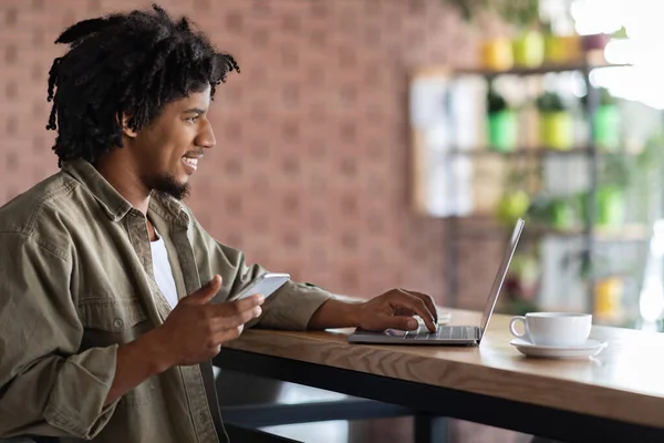 快乐的年轻的非洲卷发男子，带着智能手机在笔记本电脑上打字，坐在餐桌旁，手里拿着杯子 — 图库照片