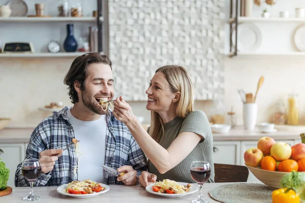 Joven mujer europea alegre alimenta a su marido de espaguetis con verduras o pasta en cocina minimalista —  Fotos de Stock