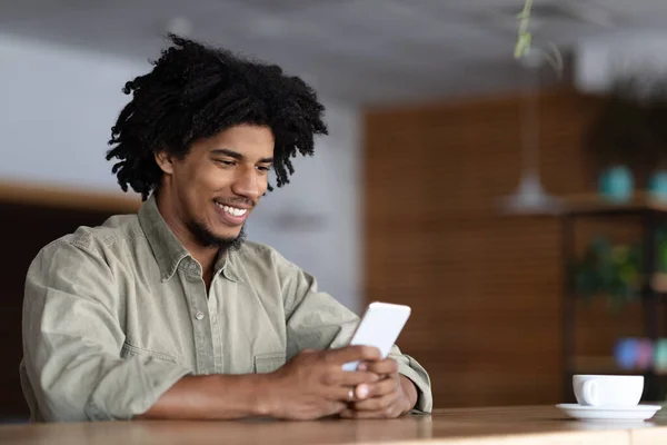 Sonriente millennial atractivo africano americano rizado hombre escribiendo en el teléfono inteligente en la mesa con café en el interior de la cafetería —  Fotos de Stock