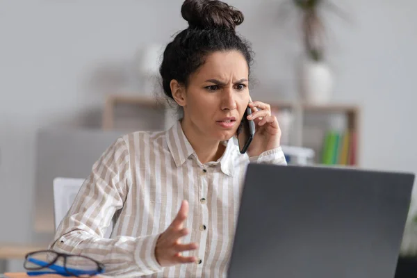 Confused young businesswoman talking on cellphone using laptop, having problem with computer, working in office — стоковое фото