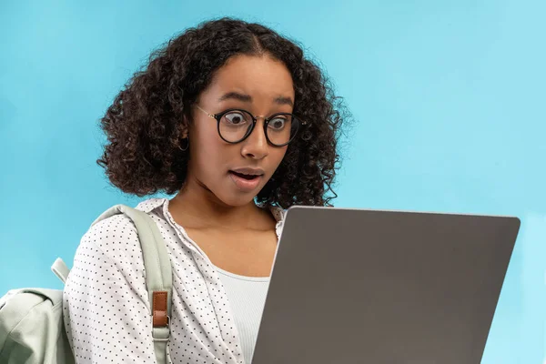 Shocked African American female student with backpack using laptop, looking at screen in surprise on blue background — Stock Photo, Image