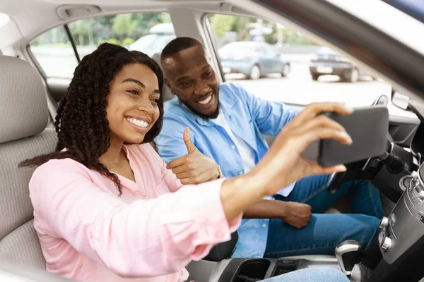 Счастливая чёрная пара водит машину и делает селфи на смартфоне — стоковое фото