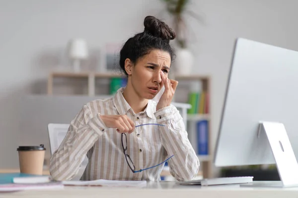 Tired unhappy female manager rubbing eyes and crying, sitting at workplace in office interior with computer — стоковое фото