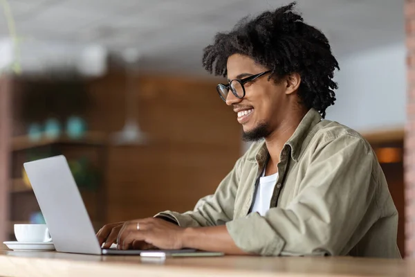 很高兴年轻的非洲卷曲的戴眼镜男性在笔记本电脑旁工作，桌上有杯子在咖啡馆里面，相貌 — 图库照片