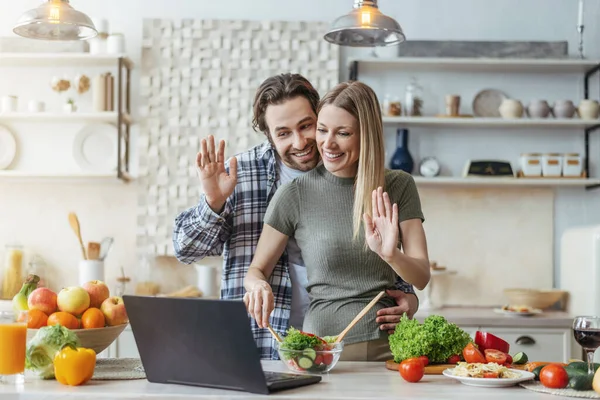 Glada unga europeiska kvinna och make med stubb förbereda sallad och viftar med händerna på laptop — Stockfoto