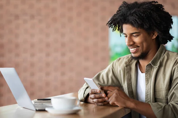 Feliz joven negro rizado chico escribiendo en el teléfono inteligente, se sienta a la mesa con el ordenador portátil, portátil y café en el interior de la cafetería —  Fotos de Stock