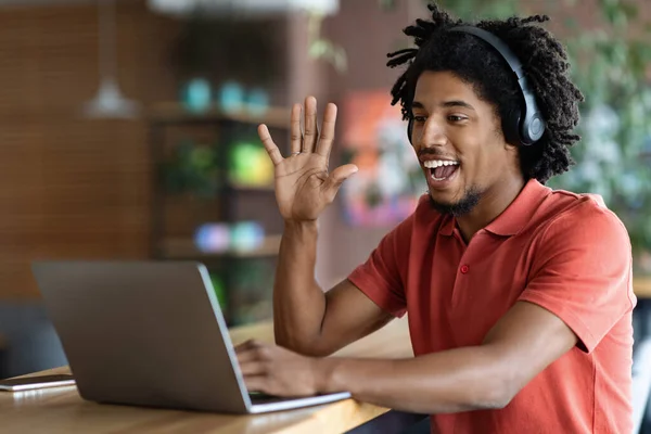 Glad young black curly guy in wireless headphones waving hand and looking at laptop at table — стоковое фото