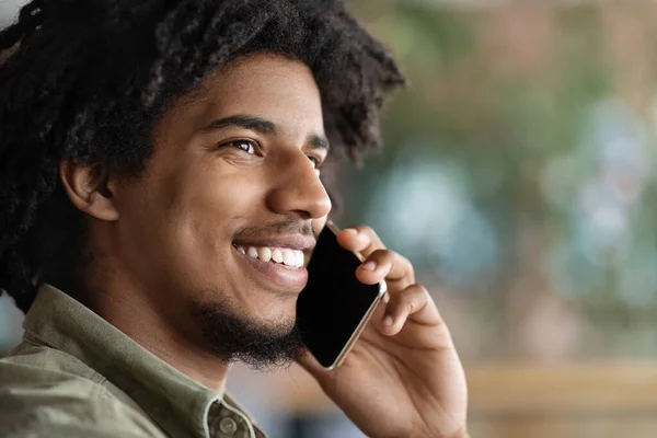Cheerful millennial african american curly guy talking by smartphone in cafe interior, close up, profile — 스톡 사진