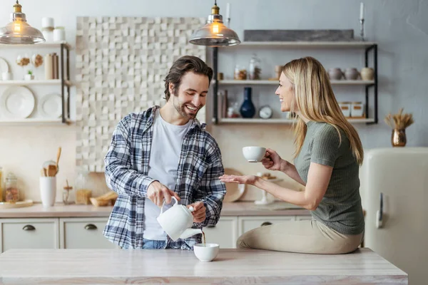 Šťastný mladý evropský manželka a manžel s strništěm pít horký čaj a mluvit ve volném čase v moderní kuchyni — Stock fotografie
