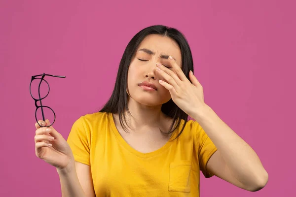 年轻的亚裔女士患有眼部疼痛，摘下眼镜，触摸着眼睛，站在紫色的背景上 — 图库照片