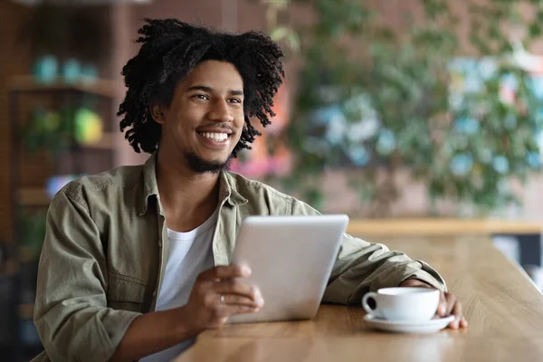 Sorrindo millennial preto encaracolado blogueiro masculino com tablet sentado à mesa, beber café e olhar para o espaço livre — Fotografia de Stock