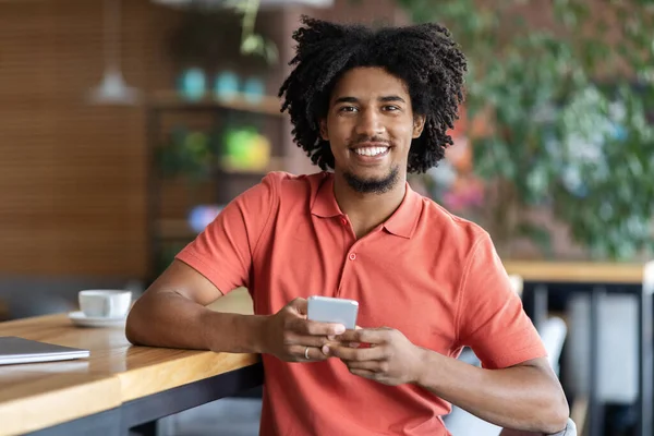 Sonriente joven afroamericano rizado mecanografía masculina en el teléfono inteligente en la mesa con taza de café en el interior de la cafetería —  Fotos de Stock