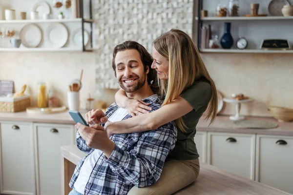 Genç Avrupalı kadın kocasına sarılıyor, adam modern mutfağın içinde akıllı telefon gösteriyor. — Stok fotoğraf
