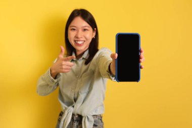 Neşeli genç Asyalı bayan boş ekranlı akıllı telefonu işaret ederek mobil uygulama, reklam sitesi, maket