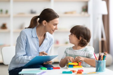 Profesyonel kadın psikolog küçük çocukla egzersiz yapıyor, okula hazır olup olmadığını kontrol ediyor ve ofiste notlar alıyor.