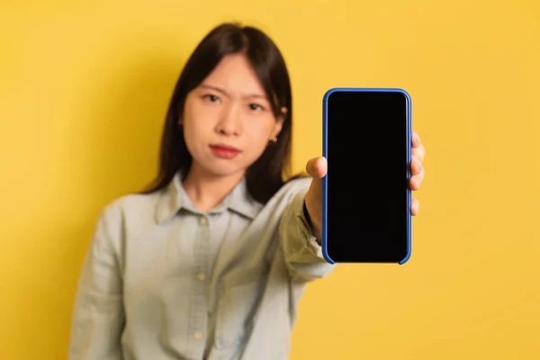 Descontente jovem mulher asiática mostrando celular com tela em branco, chateado com notícias on-line no fundo amarelo, mockup — Fotografia de Stock
