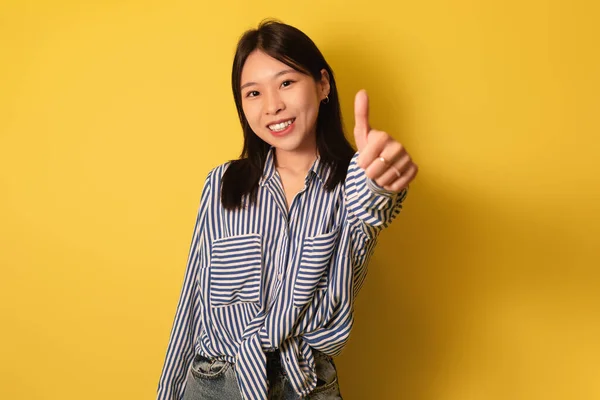 Fiatal ázsiai nő mutatja hüvelykujját felfelé, ajánló vagy jóváhagyó valamit, amely pozitív visszajelzést sárga háttér — Stock Fotó