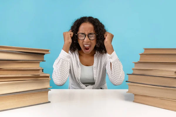Wanhopige zwarte vrouwelijke student met hopen boeken zitten aan het bureau, schreeuwen op blauwe studio achtergrond — Stockfoto