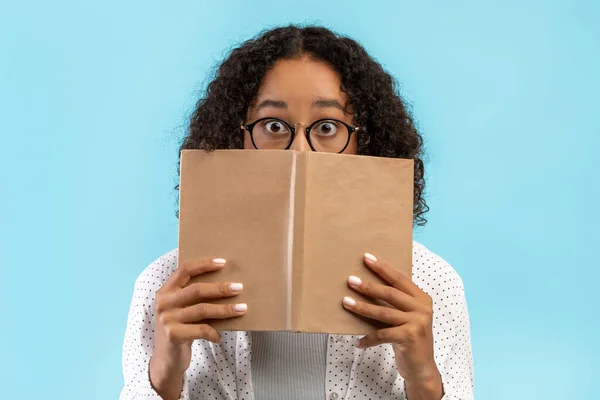 Šokovaný černošky studentka v brýlích skrývající se za knihou, studium na univerzitní zkoušky na modrém pozadí studia — Stock fotografie