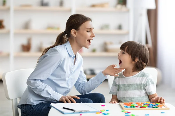 Professional woman speech therapist helping little boy to pronounce right sounds, showing mouth articulation