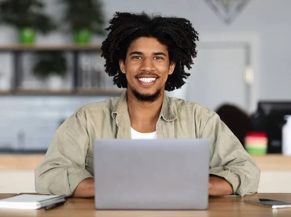 Jovem alegre bonito afro-americano encaracolado masculino trabalhando no laptop à mesa com notepad e smartphone — Fotografia de Stock