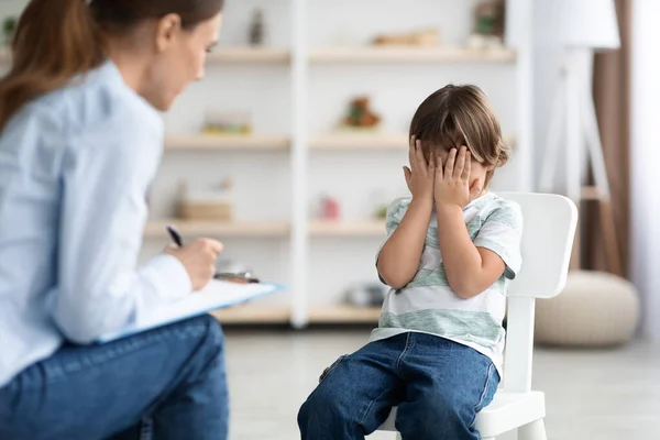 Problemas psicológicos y ayuda para los niños. Niño molesto llorando, cubriendo la cara con las manos, psicólogo tomando notas — Foto de Stock