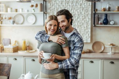Gülümseyen genç Avrupalı sakallı adam. Arkadan sarılan bayan. Çağdaş mutfakta bir fincan kahveyle.