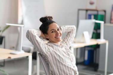 Mutlu, rahat bir kadın, elleri başının arkasında. Ofisin içinde dinleniyor, iş yerinde oturuyor.