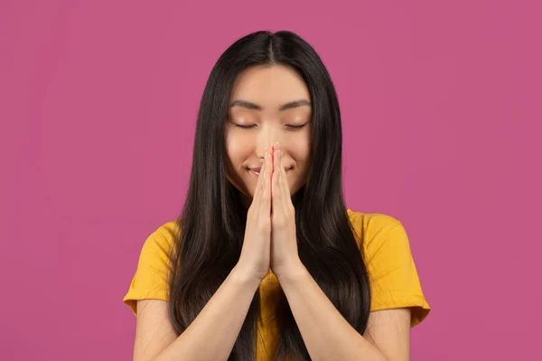 Pacífica senhora asiática orando com os olhos fechados, segurando as mãos apertadas em gesto de súplica, de pé sobre fundo violeta — Fotografia de Stock