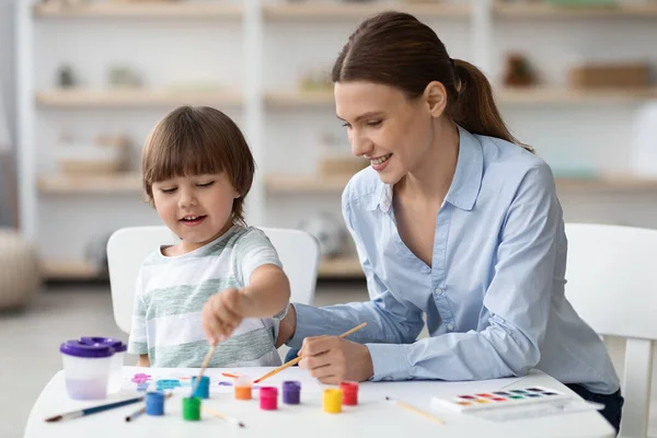 Kunstontwikkeling thuis. gelukkig jongetje schilderij met jong mooi moeder en lachen, genieten van tijdverdrijf samen — Stockfoto