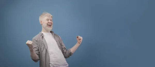 Buenas noticias. Alegre chico albino gritando y sacudiendo los puños apretados, posando sobre fondo azul, panorama — Foto de Stock