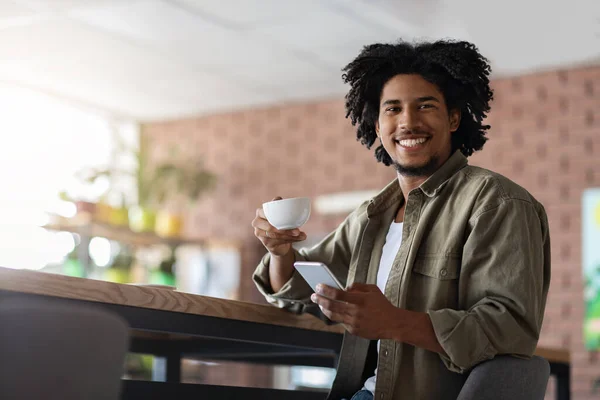 Fröhlicher Millennial afrikanisch-amerikanischer Lockenkopf mit Smartphone sitzt am Tisch, trinkt eine Tasse Kaffee — Stockfoto