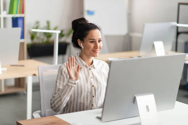 Reunión en línea. Mujer alegre haciendo videollamada en la computadora en la oficina, trabajadora de oficina en airpods saludando a la webcam —  Fotos de Stock