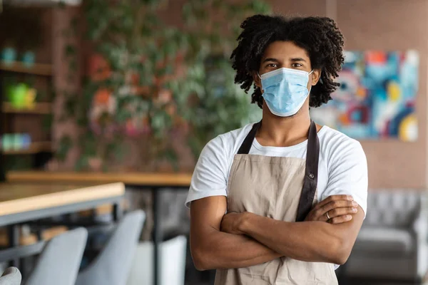 Glad confident muscular millennial african american curly man waiter in protective mask and apron with crossed hands — Stock Photo, Image